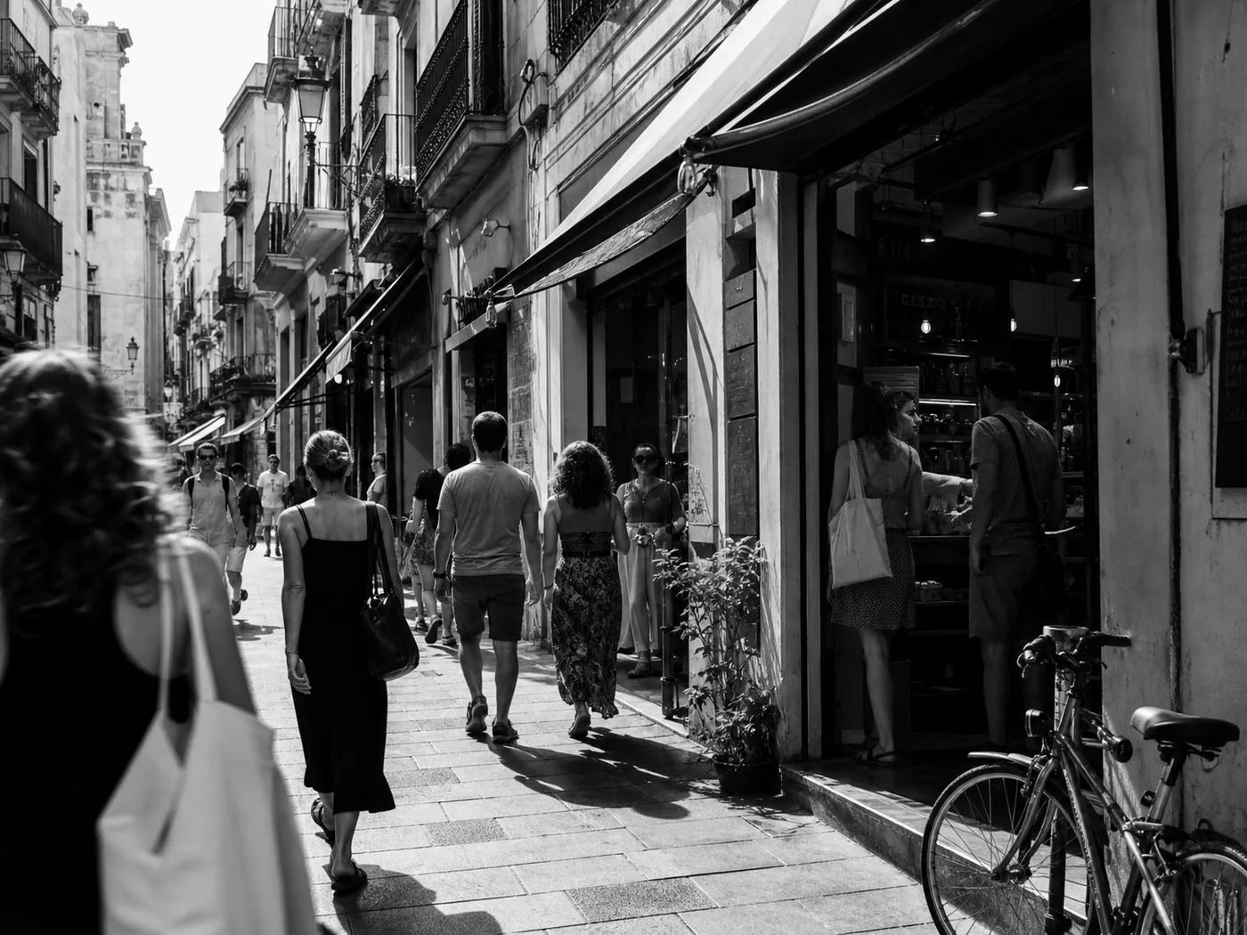 Comercios en una calle mediterránea luminosa, con personas entrando y saliendo de los locales.